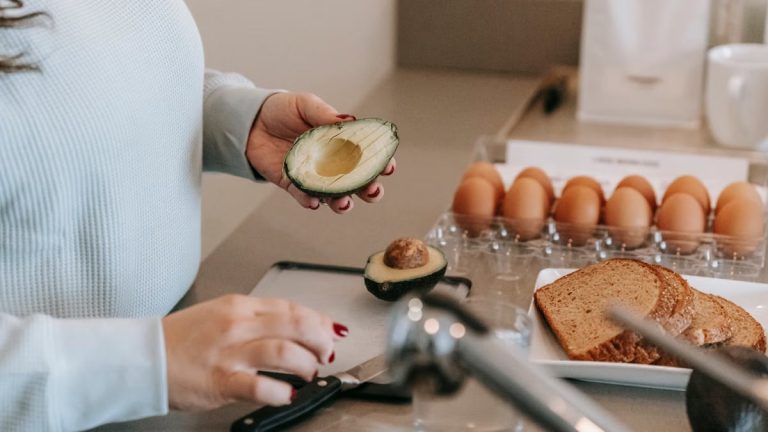 Crop faceless female cook preparing avocado while making toasts at table with sliced sliced bread and eggs in kitchen during breakfast